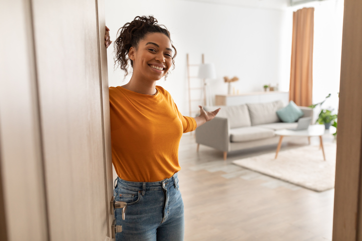 Femme afro-américaine, joyeuse et souriante, ouvrant la porte et faisant des gestes vous invitant à entrer dans son appartement